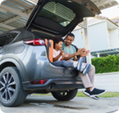 A father and his young son sit in the open hatch of an SUV parked in a garage.  